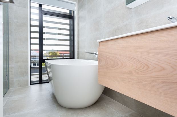 Bright contemporary bathroom featuring a freestanding white bathtub, a floating light-wood vanity with a white countertop, large pale grey stone-look wall and floor tiles, and dark-framed louvred glazing. The room has a clean, refined and airy appearance.