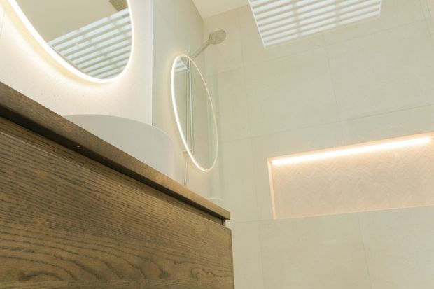 Close-up view of a contemporary bathroom vanity with a warm wood-grain drawer or cabinet front, pale tiled walls, a round illuminated mirror, and recessed linear lighting. The image emphasizes clean lines, soft warm illumination, and built-in joinery.
