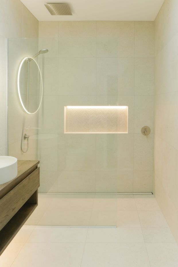 A bright, minimalist bathroom featuring a walk-in glass shower with a rainfall showerhead and handheld fixture, large pale stone-look wall and floor tiles, a recessed illuminated niche, and a floating timber vanity with a white vessel basin. The palette is warm and neutral with subtle integrated lighting.