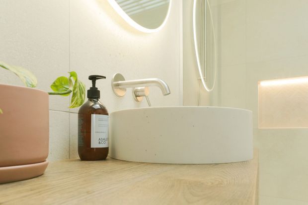 Close-up view of a refined bathroom vanity featuring a round white vessel basin on a pale timber benchtop, wall-mounted brushed metal tapware, a softly illuminated recessed wall niche, and a partially visible oval mirror. A small potted plant and soap dispenser add natural and functional details.