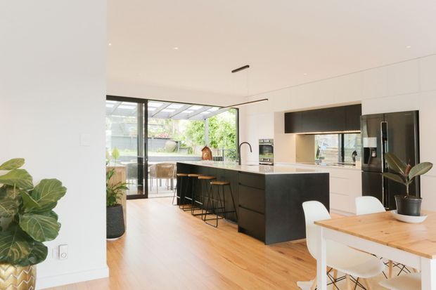 Bright contemporary open-plan kitchen and dining area with white walls and cabinetry, a dark island and integrated dark kitchen joinery, light timber flooring, a timber dining table with white chairs, and large glazed doors opening to a covered outdoor area. Indoor plants add a natural accent.