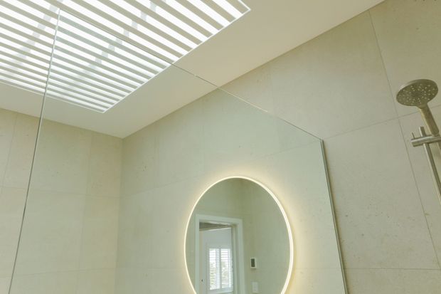 Contemporary bathroom interior featuring large pale stone-look wall panels, a round backlit mirror, a frameless glass shower enclosure, and a ceiling skylight or linear louvre opening that brings in bright natural light. A rainfall showerhead and controls are visible at right, with a doorway reflected in the mirror.