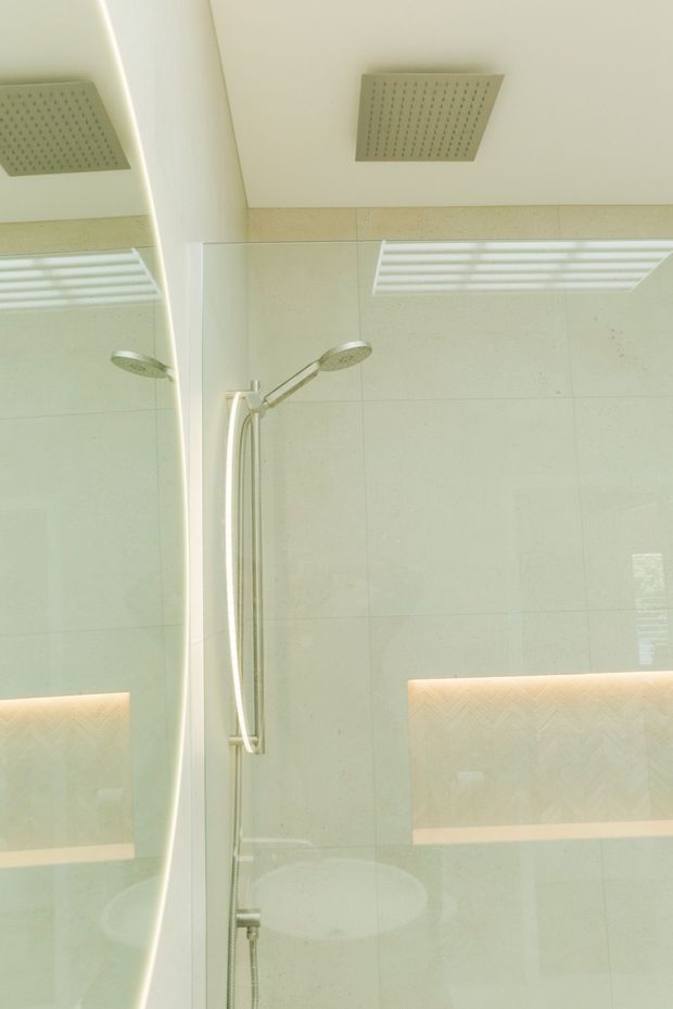 Bright contemporary bathroom shower area with pale large-format wall tiles, a frameless glass shower enclosure, twin showerheads, a handheld shower, recessed illuminated wall niches, and a large mirror reflecting the space. The palette is predominantly soft white and cream with chrome fixtures and warm niche lighting.