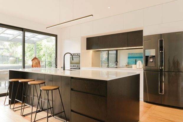 Contemporary kitchen with a large white island and integrated seating for four, dark wood-toned cabinetry, a full-height stainless steel refrigerator, mirrored splashback, and expansive black-framed glazing opening toward a bright outdoor area. Linear pendant lighting and concealed under-cabinet lighting add a clean, refined finish.