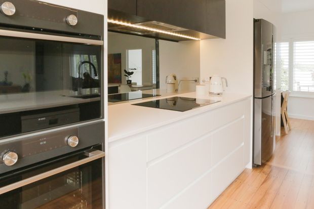 A bright contemporary galley kitchen with white handleless-style cabinetry, a white benchtop, black glass induction cooktop, integrated stainless-steel appliances, and dark overhead cupboards with under-cabinet lighting. A timber floor extends toward a small dining area by a window with white plantation shutters.