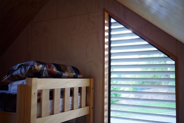 Compact timber-lined sleeping space with a wooden bunk bed and patterned bedding beside a distinctive triangular louvred window. The warm wood surfaces and angled ceiling create a simple cabin-like interior, with greenery visible beyond the window.