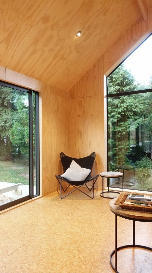 A compact, timber-lined sitting area with a dramatically sloped plywood ceiling and matching wall panels. Full-height black-framed glazing opens the corner to a leafy garden, while a black leather butterfly chair, small round side table, and round coffee table create a quiet reading or lounge nook. The warm timber surfaces and abundant natural light give the space a calm, organic character.
