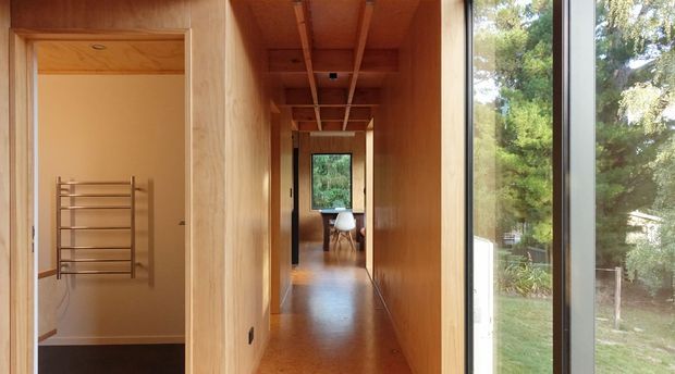 A narrow timber-lined interior hallway with a warm wood floor, exposed timber ceiling battens, and full-height glazing along one side overlooking greenery. A bathroom with a wall-mounted heated towel rail opens directly off the hall, while a dining or living area is visible at the far end.