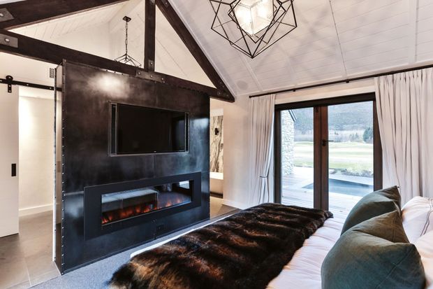 A cozy bedroom with a low bed, layered white bedding, dark fur throw, and large cushions. A dramatic black steel feature partition incorporates a television and built-in electric fireplace, while exposed dark timber beams, a white-lined pitched ceiling, geometric pendant lighting, and full-height glazed doors create a warm contemporary lodge aesthetic. The glazing opens toward a deck and landscaped exterior.
