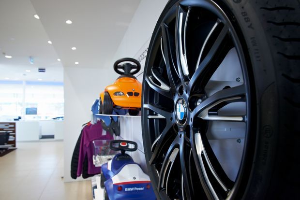 Close-up view of a large black and silver BMW alloy wheel and tire displayed prominently in a bright, modern showroom. Behind it are branded automotive products and accessories, including a child-sized ride-on vehicle and clothing, with white cabinetry, polished light flooring, recessed ceiling lights, and glazed frontage visible in the background.