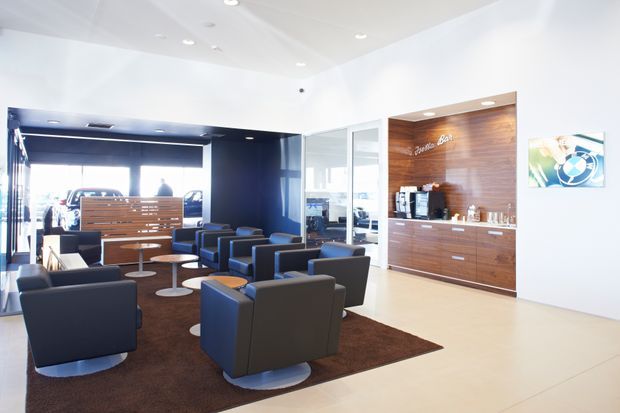 Bright contemporary commercial lounge and reception area with multiple dark upholstered lounge chairs arranged on a large brown rug. A timber reception or beverage counter with integrated cabinetry and a small illuminated sign is set against a warm wood feature wall, with a glossy dark partition, glass panels, and large glazing contributing to the open layout. Recessed ceiling lighting and pale flooring create a clean, professional atmosphere.