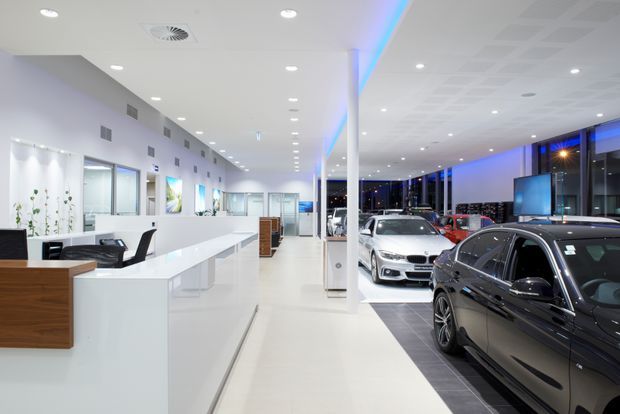Bright, contemporary car dealership showroom with a long white reception and sales counter in the foreground, several vehicles displayed on dark tiled flooring, extensive glazing, recessed ceiling lights, and blue accent lighting along the ceiling and display areas.