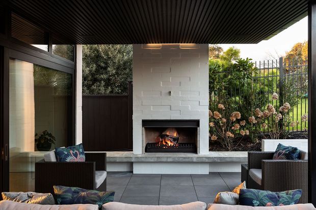 A covered outdoor lounge or terrace with a built-in fireplace as the focal point. The space features a dark slatted ceiling, glazed wall and doors, a white painted brick fireplace surround, a long grey stone or concrete hearth, large-format grey tiled flooring, and dark woven outdoor seating with patterned cushions. A planted garden is visible beyond the metal fence.