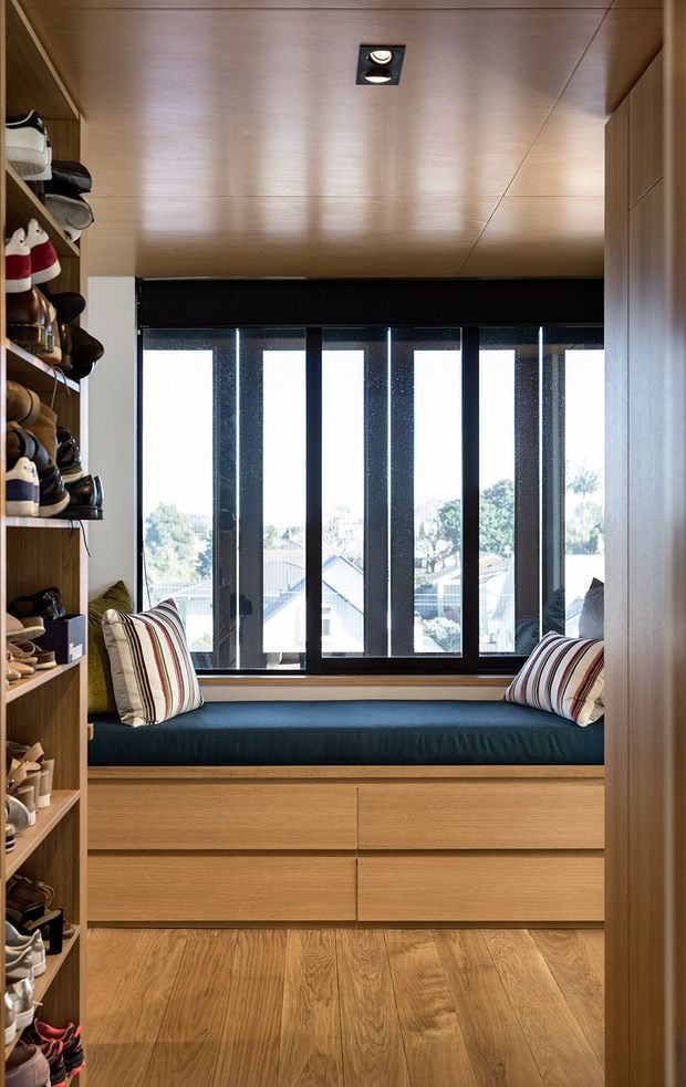 A compact built-in window seat occupies the end of a narrow dressing or wardrobe space. Full-height open shelving on the left displays shoes, while extensive light timber cabinetry surrounds the seat and continues beneath it as drawers. The seat has a dark teal cushion and striped cushions, with a large black-framed window overlooking a suburban neighbourhood. The ceiling is clad in warm timber panels and includes a recessed downlight; the floor is also timber.