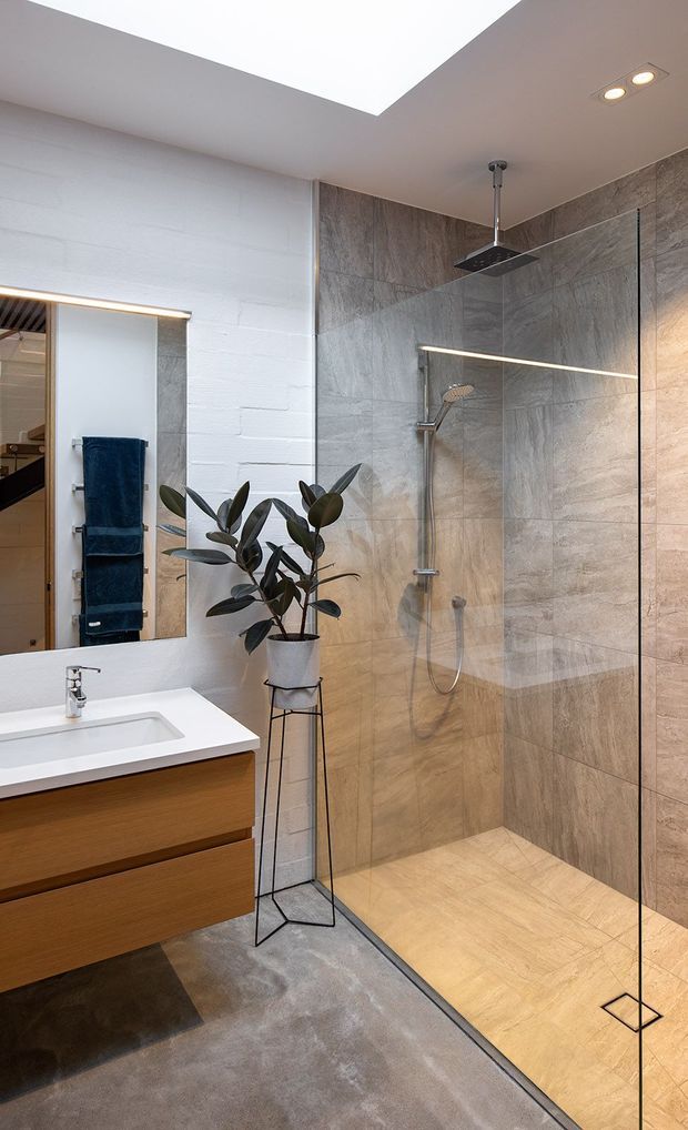 A contemporary bathroom featuring a walk-in glass-enclosed shower with large grey-beige stone-look wall and floor tiles, a wall-mounted timber vanity with white integrated basin, and a large frameless mirror. A potted rubber plant adds greenery, while recessed and linear lighting create a bright, refined atmosphere. The ceiling includes a large rectangular skylight or lightwell.