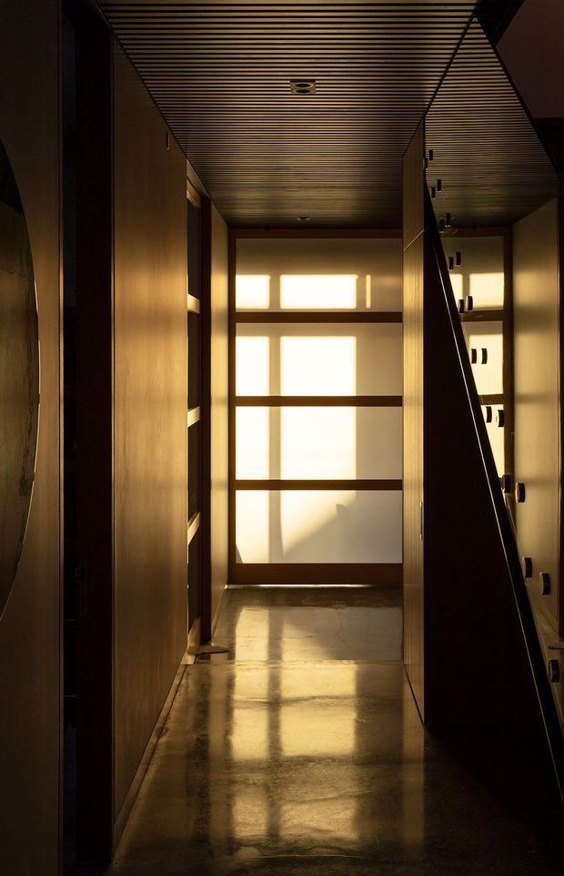 A narrow, dimly lit interior passage with warm timber-toned built-in cabinetry and a slatted timber ceiling. A large frosted or translucent window at the end filters strong golden sunlight into the corridor, reflecting across the polished floor. Dark shelving or joinery lines the right side, creating a dramatic, intimate atmosphere.