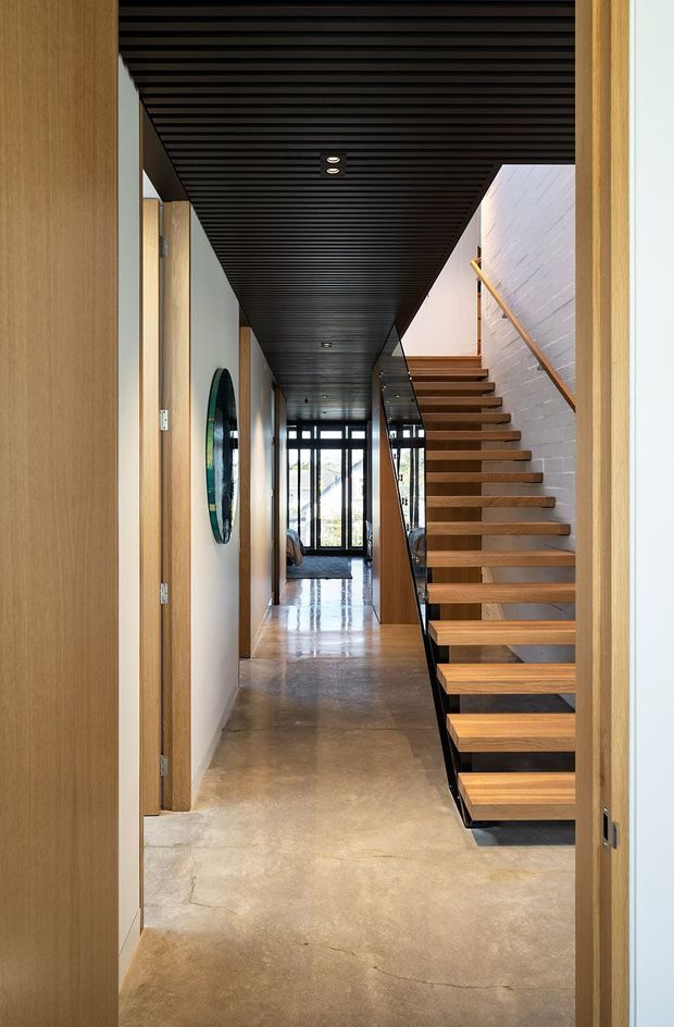 A contemporary residential hallway viewed through a timber-framed doorway, featuring a floating open-riser timber staircase with black steel supports, a polished concrete floor, white-painted brick stair wall, and a dramatic black linear-slat ceiling with recessed downlights. Full-height glazing is visible at the end of the corridor, while a round wall artwork and timber doors add detail.