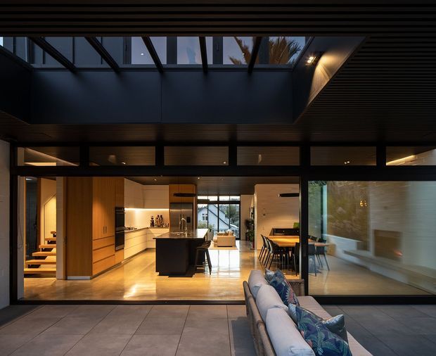 A contemporary open-plan home viewed from an outdoor terrace through extensive floor-to-ceiling glazing. The interior features a warm timber-lined kitchen with a central island, integrated cabinetry and appliances, an adjacent dining area, and a bright living space. Strong black structural framing, glazed clerestory panels and a skylight-like roof opening create a dramatic indoor-outdoor connection. A large outdoor sofa occupies the foreground.