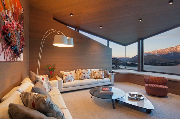 A warm, contemporary living room with extensive timber wall and ceiling lining, a large cream sectional sofa, sculptural arc floor lamp, low geometric coffee tables, and colourful artwork and cushions. Full-height corner glazing frames a dramatic mountain and lakeside landscape.