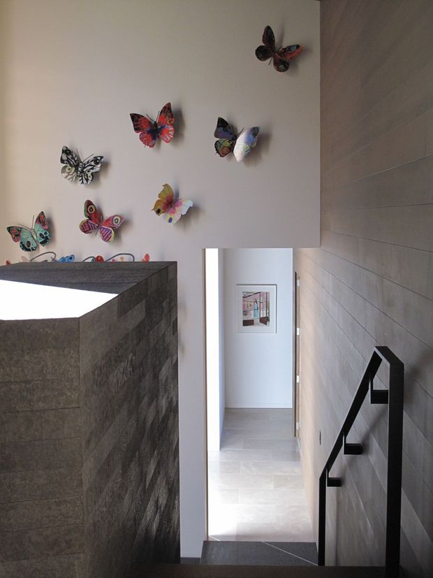 A narrow contemporary hallway and stairway with a tall void. The right wall is clad in horizontal light-grey panels or tiles and has a simple black angular handrail. A dark stone-clad stair or partition occupies the left foreground, while a brightly lit corridor extends toward a framed artwork. Colourful butterfly wall decorations add a playful accent to the otherwise restrained space.