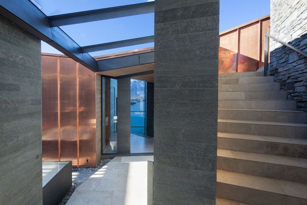 A modern architectural entry courtyard or circulation space with exposed dark masonry walls, broad concrete stairs, copper-toned metal screening, and a glazed roof canopy. A glass doorway leads into the building, while a stacked-stone wall lines the stair edge. The palette is dark grey, copper, and cool blue, with strong geometric forms and natural daylight.