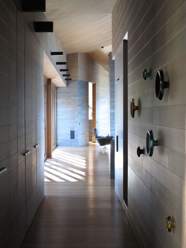 A long, narrow contemporary hallway with dark grey tiled wall panels, warm timber flooring and a timber-lined ceiling. Linear black wall-mounted lights run along the left side, while the right wall features sculptural circular controls or decorative fittings. At the end, the corridor opens toward a brighter sitting area with a curved grey stone-clad wall, timber doors, and a black lounge chair. Strong sunlight creates striped shadows across the floor.