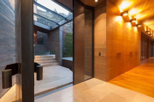A refined entry corridor with warm timber wall and ceiling panelling, integrated lighting, dark-framed full-height glazing, and a large glass roof opening to a landscaped external stairwell. Pale stone or tile flooring contrasts with the rich wood and dark metal detailing.