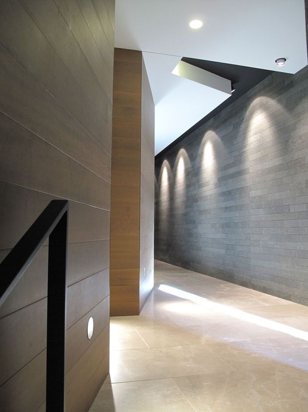 A dramatic contemporary interior passage or stair landing with a tall void, large-format pale stone or porcelain floor tiles, warm wood-toned wall panelling, and a dark grey horizontally coursed stone feature wall. Recessed spotlights create repeated pools of light along the feature wall, while a sharp angular ceiling opening or skylight brings in daylight. A black stair balustrade is visible in the foreground.