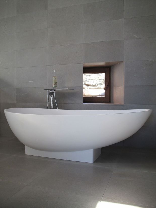 A minimalist bathroom featuring a large freestanding white oval bathtub on a rectangular plinth, set against full-height grey tiled walls and a matching tiled floor. A wall-mounted bath tap and handheld shower sit behind the tub, beside a small timber-framed recessed window.