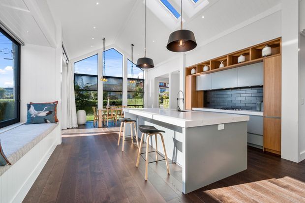 Bright contemporary open-plan kitchen with a large white island, integrated seating, dark timber flooring, and timber-framed cabinetry with open display shelving. A dark tiled splashback, pendant lighting, clerestory glazing, and a built-in window seat define the space, with a dining and outdoor area visible beyond the glazing.
