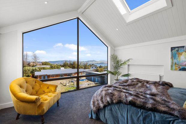Bright contemporary bedroom with a dramatic angled floor-to-ceiling corner window overlooking a suburban landscape and distant hills. The room has a pitched, white-lined ceiling with skylights, dark carpet, a blue upholstered bed covered with a textured fur throw, a mustard-yellow armchair, a tall indoor palm, and colourful wall art.