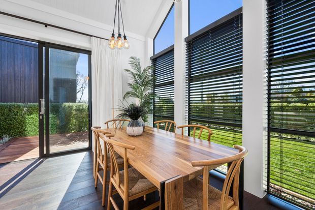 Bright contemporary dining area with a large solid timber dining table and matching timber chairs, set beside full-height glazing and dark horizontal venetian blinds. A glass door opens to a landscaped garden, while pendant lights and a potted palm add warmth and texture. The room has a high, light-coloured ceiling and dark timber-look flooring.