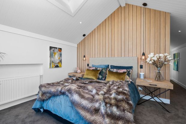 A bright, contemporary bedroom with a large bed dressed in blue bedding, patterned cushions, and a textured throw. The feature wall combines vertical light timber panelling with a grey upholstered headboard, while matching pendant lights hang beside the bed. White walls and a pitched, white-lined ceiling create an airy setting, complemented by a timber bedside table, artwork, radiator, and dark carpet.