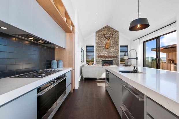 Bright, open-plan contemporary kitchen overlooking a living area. The kitchen features white stone benchtops, dark grey cabinetry, a dark tiled splashback, integrated appliances, timber open shelving, and a large island with sink. Beyond it is a high-ceiling living room with a prominent exposed brick fireplace and glazing opening to an outdoor deck.
