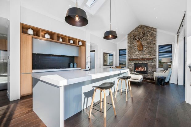 Open-plan contemporary kitchen and living area with a large white island and breakfast bar, pale blue-grey cabinetry, warm timber open shelving, dark tiled splashback, and dark timber flooring. The living zone features a tall exposed-brick fireplace wall with a mounted deer head, skylights, pendant lighting, and high ceilings.