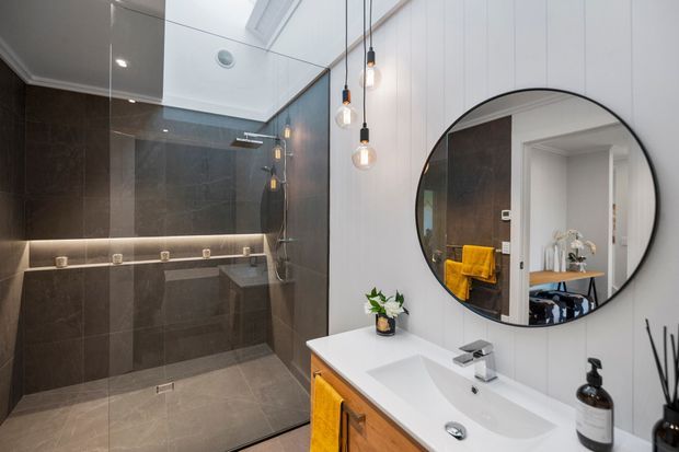 Contemporary bathroom featuring a large walk-in glass shower lined with dark grey stone-look tiles, a floating timber vanity with a white integrated basin, round black-framed mirror, and pendant lighting. A skylight or roof window brings natural light into the space, while yellow towels provide a warm accent.