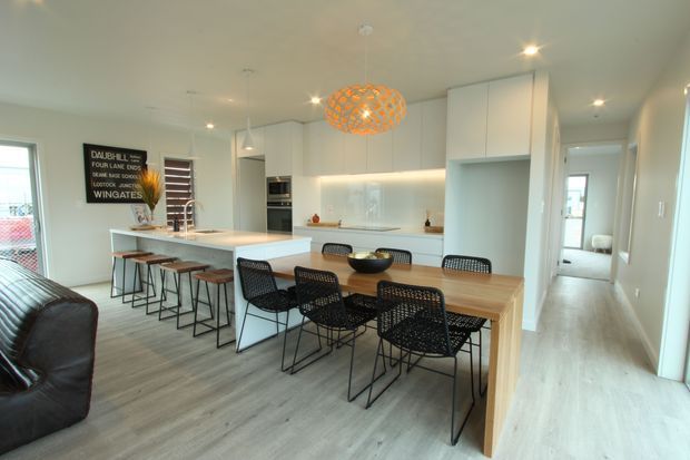 Open-plan kitchen and dining area with white floor-to-ceiling cabinetry, a large pale benchtop island with bar seating, and a timber dining table surrounded by black woven chairs. Light wood-look flooring, recessed ceiling lights, pendant lighting, and a bright neutral palette create a clean contemporary interior. A living area and hallway are visible beyond the main space.