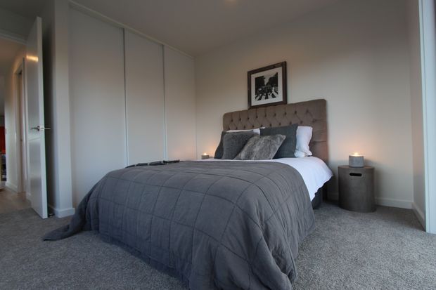 A calm, contemporary bedroom with a large upholstered button-tufted headboard, layered grey bedding, white pillows, built-in white wardrobes, grey carpet, and a dark bedside table with a small lamp. The room has neutral finishes, soft lighting, and a framed monochrome artwork above the bed.