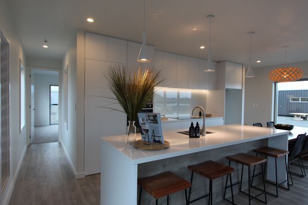 Contemporary open-plan kitchen with a large white island and integrated cabinetry, fitted with timber-topped bar stools, pendant lighting, and a stainless-steel faucet. A hallway leads toward the rear of the home, while glazing opens the kitchen toward an exterior deck or courtyard.