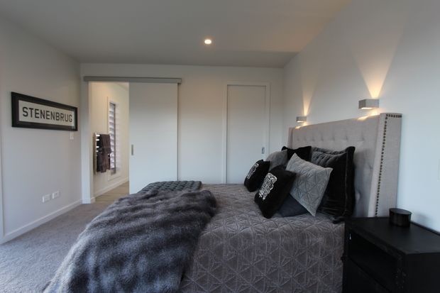 A modern, softly lit bedroom featuring a large upholstered bed with a tall grey headboard, grey patterned bedding, dark cushions, and a textured throw. White walls, recessed ceiling lighting, integrated bedside wall lights, carpeted flooring, and simple sliding wardrobe doors create a clean, contemporary interior.