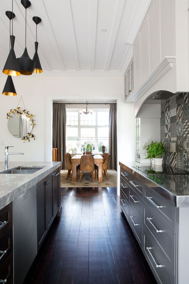 A bright galley-style kitchen leading into a dining area. The kitchen features dark painted cabinetry, pale stone benchtops, a patterned geometric tile splashback, white decorative wall panelling and a coffered or panelled ceiling. Matte black pendant lights with warm metallic interiors hang above the island, while dark timber flooring runs through to the dining space. A large window provides abundant natural light.