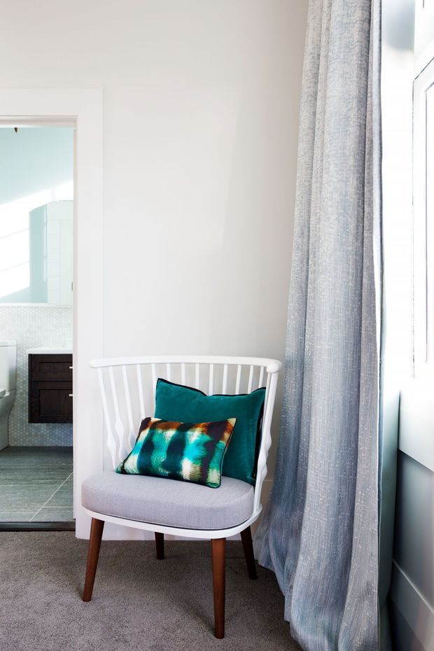 Bright interior corner featuring a white spindle-back accent chair with tapered timber legs, grey upholstery, and teal patterned cushions beside a large window with sheer grey curtains. A tiled bathroom with a dark timber vanity is visible through the open doorway.
