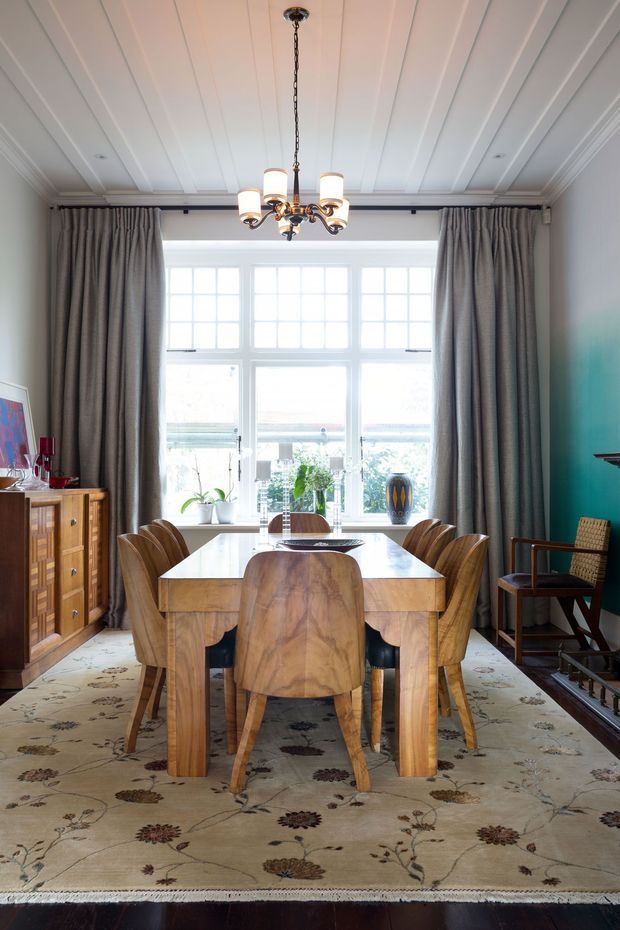 A refined dining room with a large timber dining table and matching sculptural wooden chairs, positioned in front of a bright multi-pane bay window. The room features a white panelled and coffered ceiling, full-height grey curtains, a floral patterned rug, a traditional timber sideboard, teal painted walls, and a brass-and-glass chandelier. The overall look combines traditional architectural detailing with richly crafted mid-century-inspired furniture.