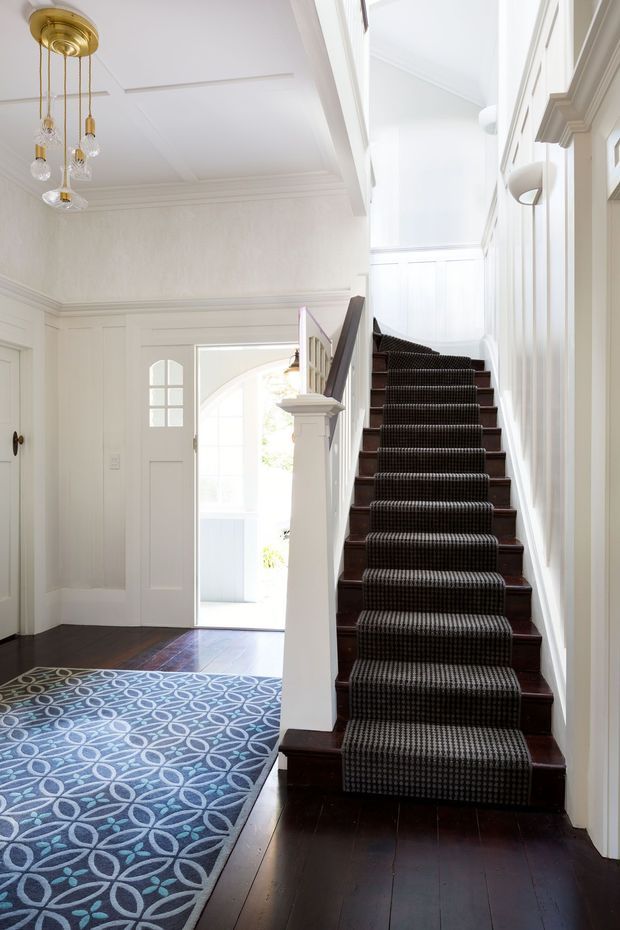 Bright, elegant residential entry hall with a dark timber staircase and patterned runner rising beside white painted wall panelling. The space features a high ceiling with decorative boxed moulding, traditional trim and wainscoting, a white paneled front door, and a brass multi-light pendant. A blue geometric rug extends through the entry, while daylight enters through the open doorway and tall stairwell glazing.
