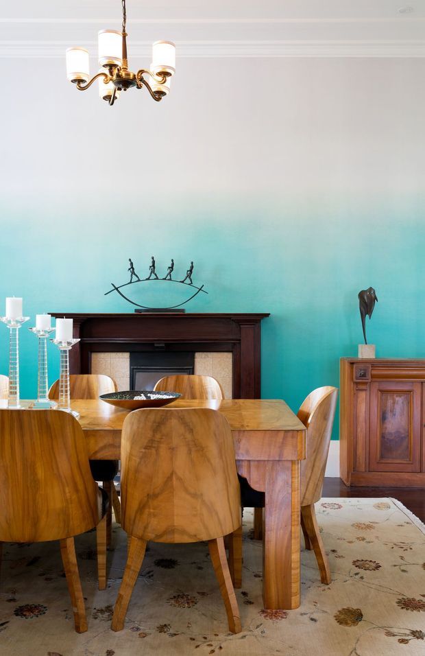 A bright traditional dining room with a large polished timber dining table and matching curved-back timber chairs. The room features a dark wood fireplace mantel with tiled surround, a turquoise-to-white painted feature wall, ornate white cornices, a brass chandelier, a timber sideboard, decorative candlesticks, and a patterned area rug.