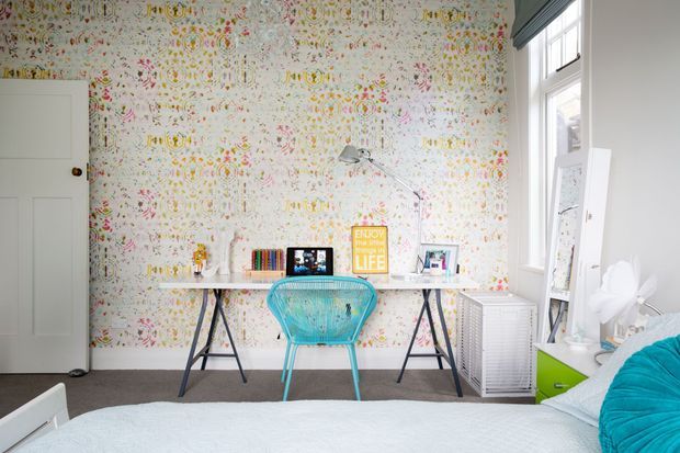 Bright bedroom workspace featuring a white trestle desk, turquoise chair, patterned floral wallpaper, white-painted door and trim, tall window, and colorful bedside furnishings. The bed is visible in the foreground, confirming the room as a bedroom with a compact home office area.