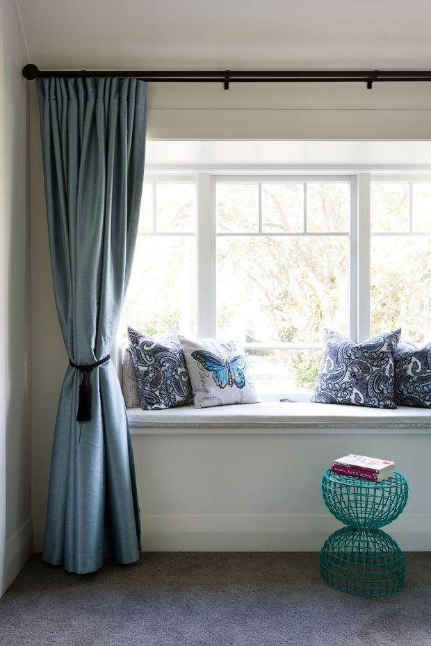 A bright residential window seat beneath a large multi-pane window, framed by full-length blue-grey curtains on a dark rod. The built-in sill is styled with patterned blue-and-white cushions, including a butterfly motif, while a turquoise wire side table or stool holds books. The room has pale walls, grey carpet, and abundant natural light.