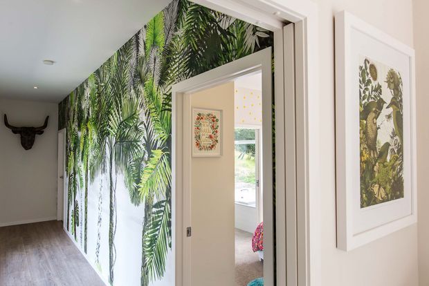 Bright residential hallway with pale walls, light timber flooring, white door and window trims, and a dramatic floor-to-ceiling tropical botanical mural on the central wall. Framed botanical artwork and open doorways lead toward a naturally lit room beyond.