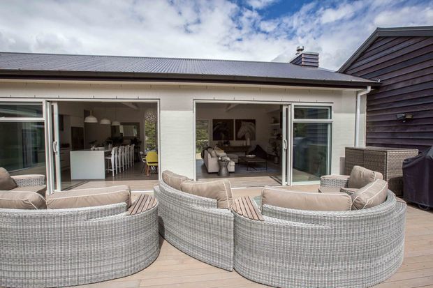 A contemporary outdoor deck or patio viewed from the garden, furnished with large curved woven outdoor lounge seating and cushions. Wide sliding glass doors open into a bright interior combining a kitchen and living area, while dark horizontal cladding forms part of the adjacent exterior wall. The home has a low-pitched metal roof and a neutral, relaxed palette.