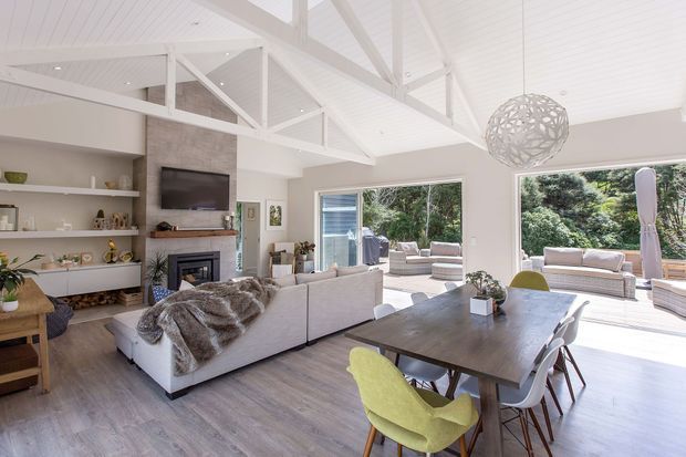 Bright open-plan living and dining area with a white vaulted ceiling, exposed painted roof trusses, large glazing opening to an outdoor seating area, a stone fireplace wall with built-in shelving and television, a large dining table, and a neutral sectional sofa. A sculptural pendant light and greenery add decorative accents.